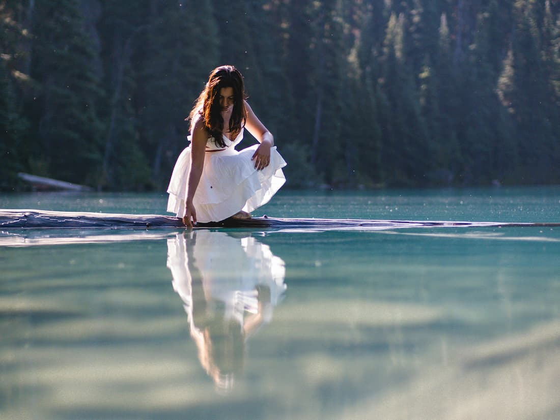 Frau in weißem Kleid berührt still die Wasseroberfläche eines Sees – Symbol für Yin Wasser und sanfte Selbstverbindung im Juli.